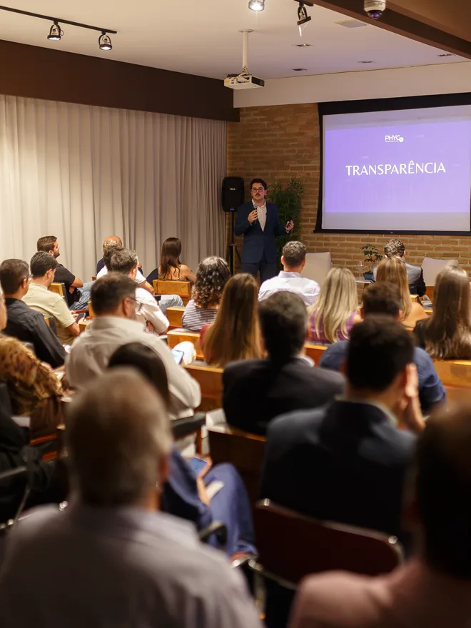 Gabriel Madona apresentando para uma plateia cheia, com a palavra Transparência projetada na tela