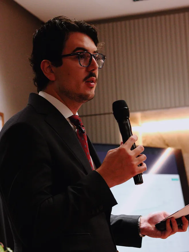 Gabriel Madona apresentando ao microfone, com anotações na mão e um gráfico projetado ao fundo